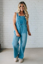 Woman wearing blue denim overalls standing against a white brick wall.