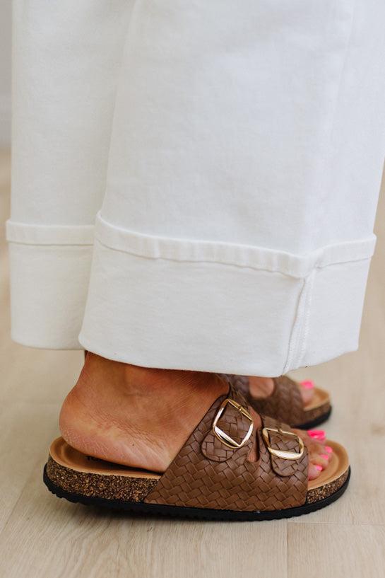 Brown sandals with gold buckles worn with white pants on a light wooden floor.