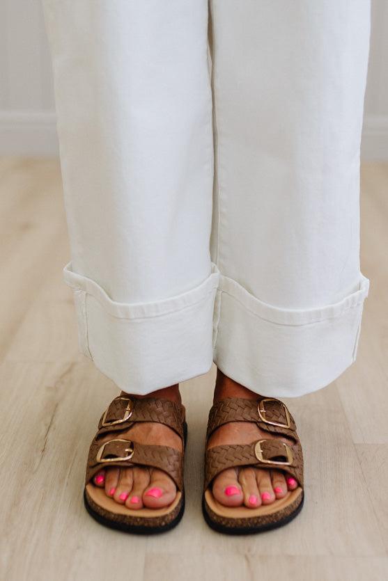 Brown sandals worn with white pants on a light wooden floor.