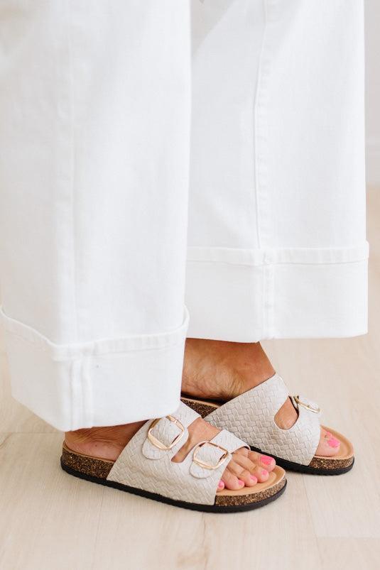 White sandals with gold buckles worn by a person with pink nail polish on a light wooden floor.