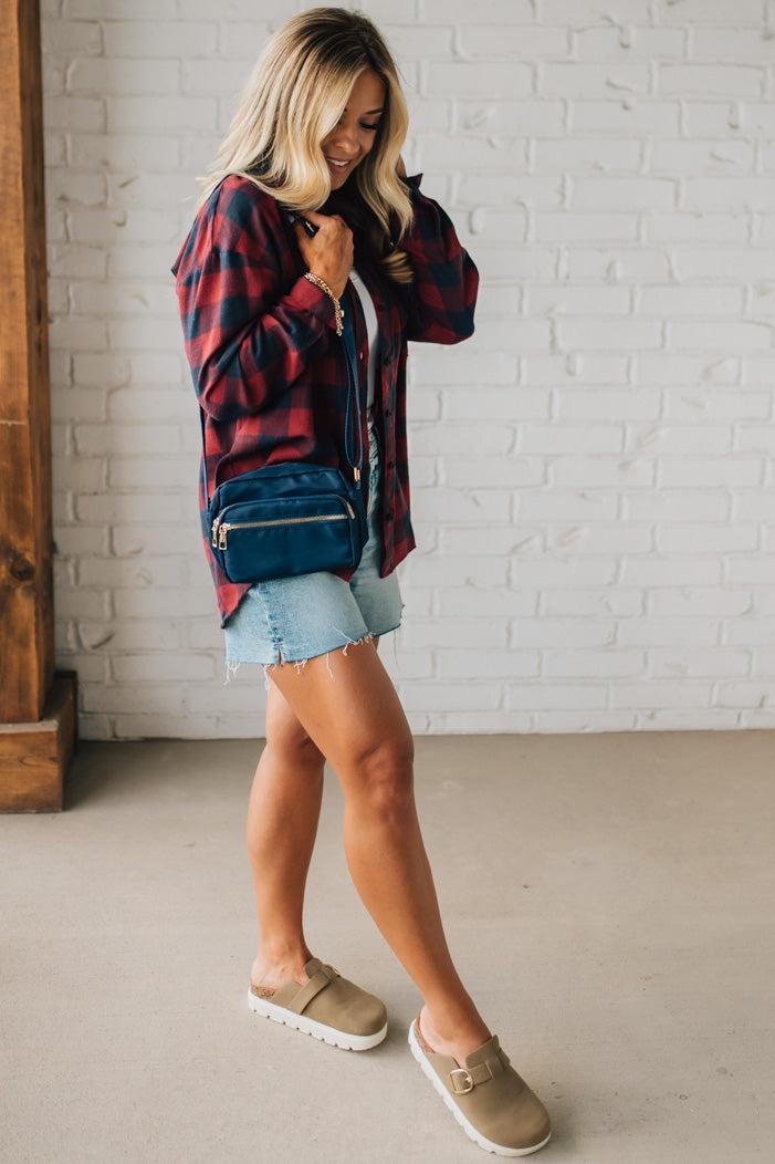Woman wearing a red plaid shirt, denim shorts, and beige sneakers against a white brick wall.