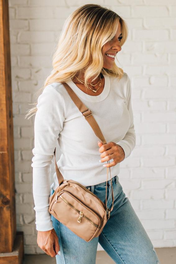 Woman holding a beige crossbody bag against a white brick wall.