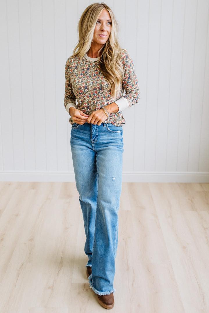 A woman modeling a long-sleeve crewneck sweater with a dense multicolor floral pattern, paired with light-wash blue jeans and brown boots, standing in a room with light wood floors and white paneled walls.