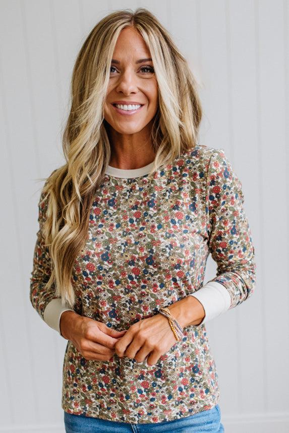 A smiling blonde woman wearing a long-sleeve crewneck top with a dense multicolor floral print on a beige background, paired with blue jeans, set against a white paneled wall.
