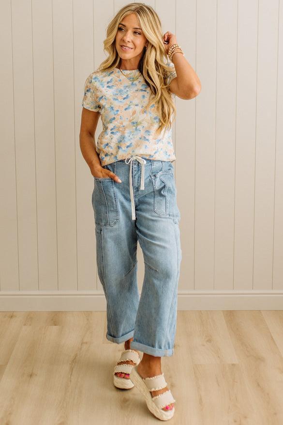 Woman wearing a floral top and light blue jeans standing against a white paneled wall.