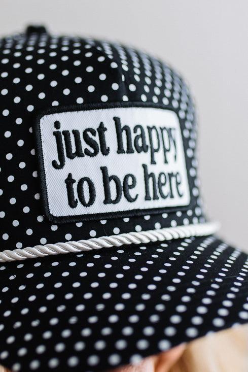 BLONDE WOMAN WEARING A BLACK AND WHITE POLKA DOT HAT WITH WHITE ROPE ACROSS THE FRONT AND A PATCH THAT SAYS JUST HAPPY TO BE HERE ON THE CENTER FRONT.