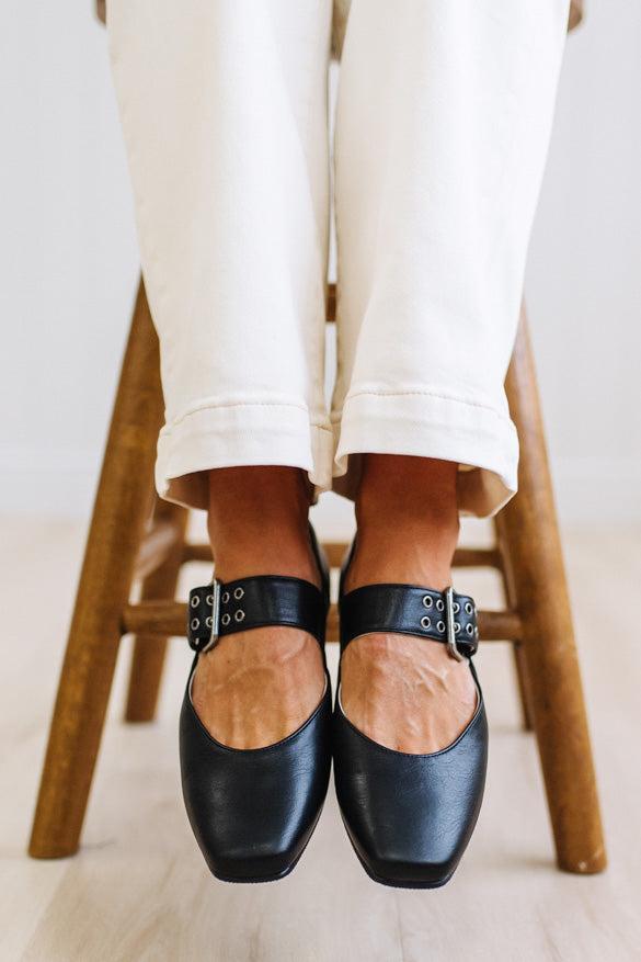 A person wearing black leather Mary Jane flats with silver-tone buckle straps, paired with off-white cuffed pants, sitting on a wooden stool.