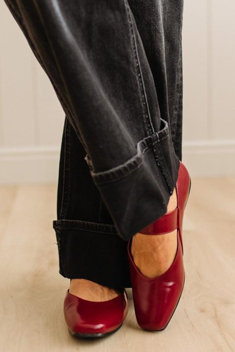 Red shoes worn with black pants on a light wooden floor.