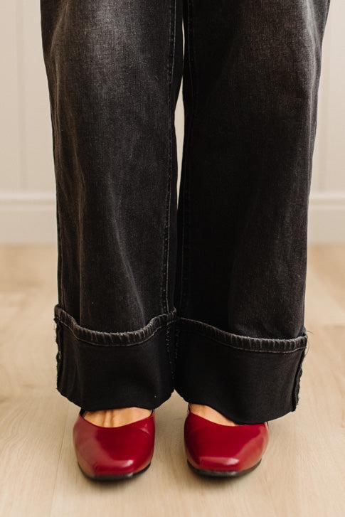 Black wide-leg jeans with rolled-up cuffs worn with red shoes on a light wooden floor.