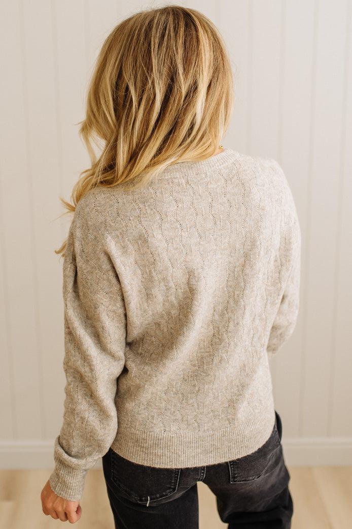 Person wearing a beige sweater and dark pants against a white wall.