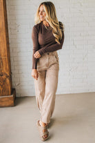 Woman wearing a brown long-sleeve top and beige pants standing against a white brick wall.
