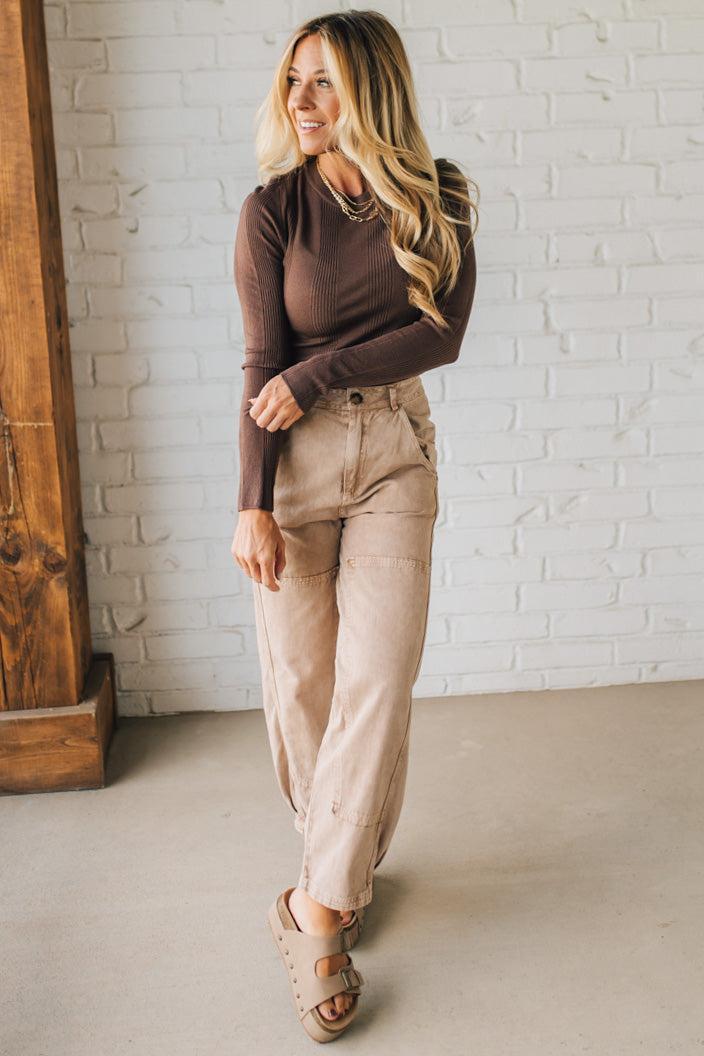 Woman wearing a brown long-sleeve top and beige pants standing against a white brick wall.