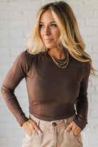 Woman wearing a brown ribbed sweater and beige pants against a white brick wall.