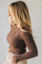 Woman wearing a brown ribbed long-sleeve top against a white brick wall.