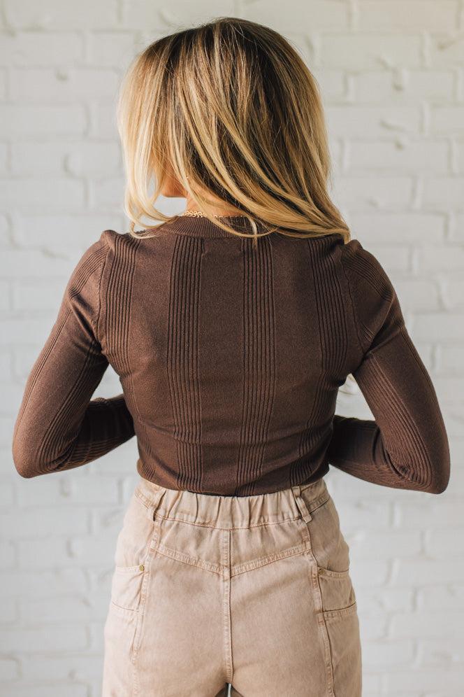 Person wearing a brown ribbed long-sleeve top and beige pants against a white brick wall.