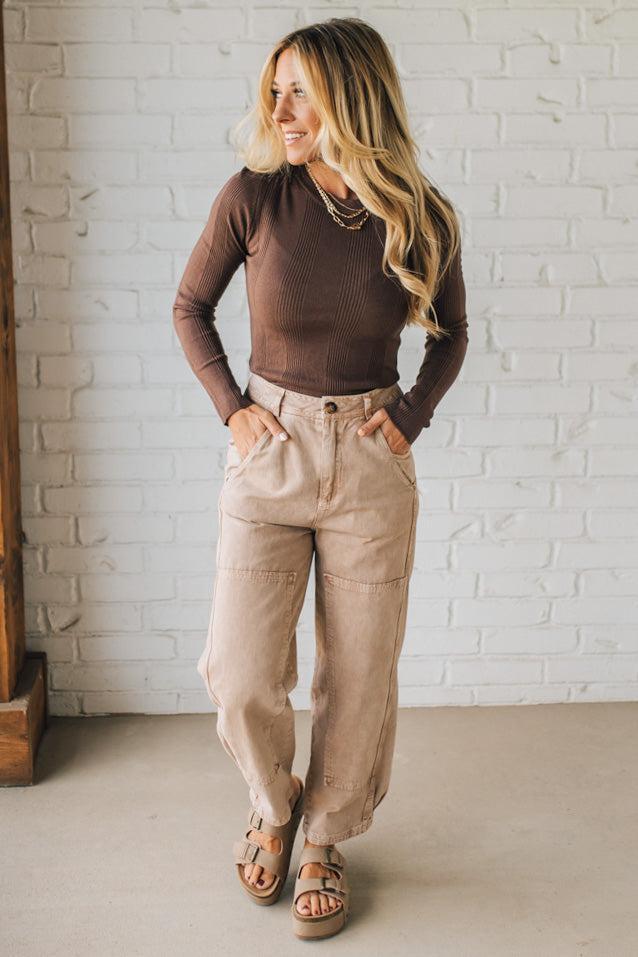Woman wearing a brown top and beige pants standing against a white brick wall.