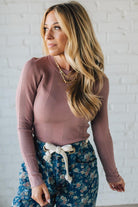 Woman wearing a mauve sweater and floral pants against a white brick wall.
