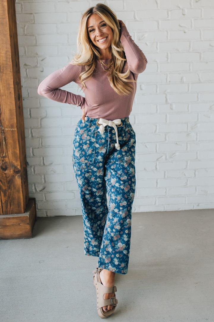 Woman wearing a pink long-sleeve top and blue floral pants against a white brick wall.