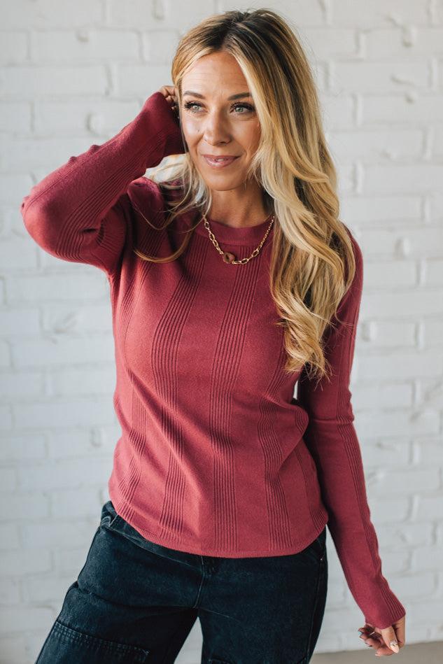 Woman wearing a red sweater and dark jeans against a white brick wall.