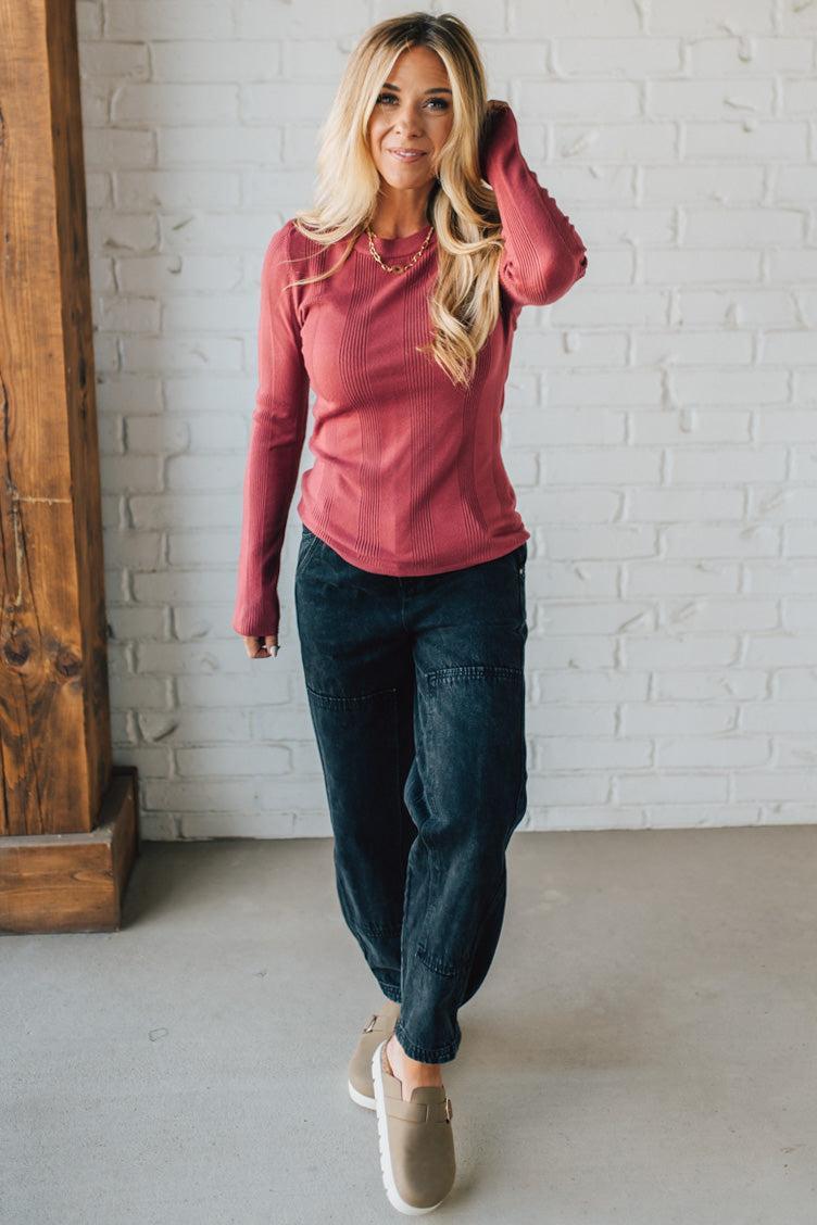Woman wearing a pink long-sleeve top and dark blue pants against a white brick wall.