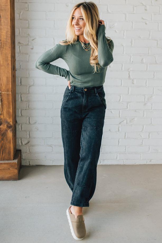 Woman wearing a green sweater and dark jeans standing against a white brick wall.