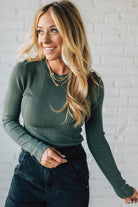 Woman wearing a green ribbed sweater and dark pants against a white brick wall.