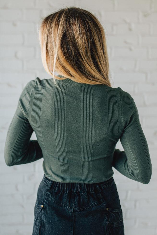 Person wearing a green ribbed sweater against a white brick wall