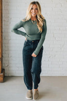 Woman wearing a green long-sleeve top and dark jeans standing against a white brick wall.
