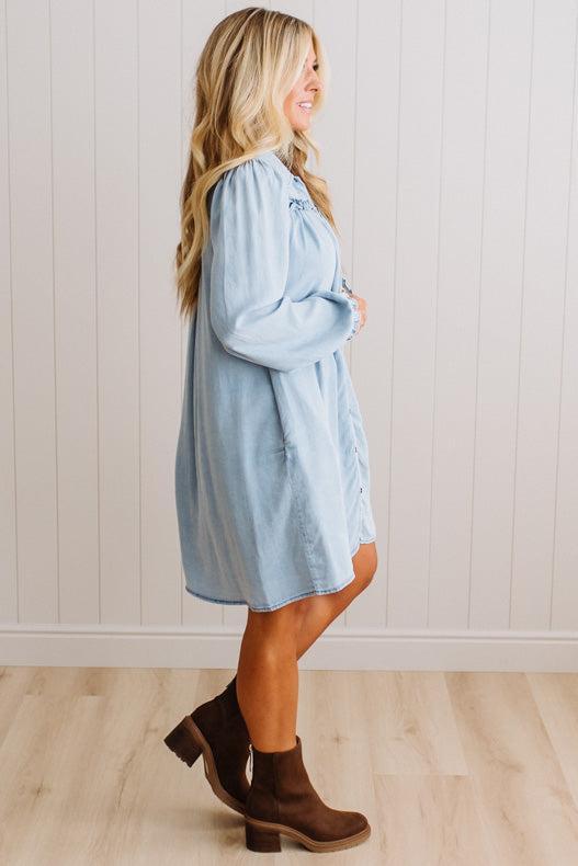 A woman modeling a light blue denim shirt dress with brown ankle boots, standing in profile against a white paneled wall and light wood floor.