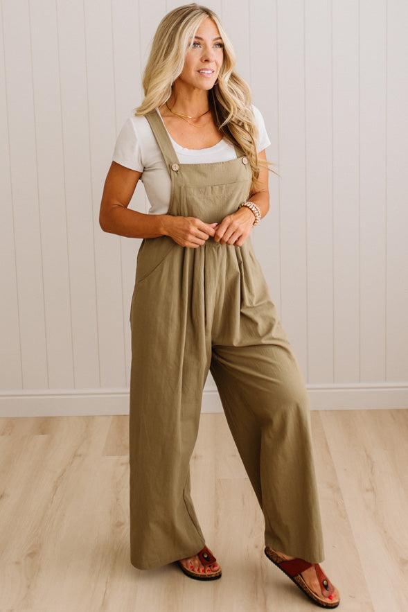 A woman modeling wide-leg utility jumpsuit in a light khaki color, worn over a white t-shirt and brown sandals, standing in front of a white paneled wall and light wood floor.