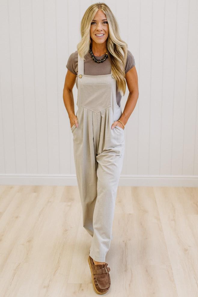 A woman modeling a pair of wide-leg stone colored utility jumpsuit over a brown t-shirt, standing in a room with light wood floors and white paneled walls.