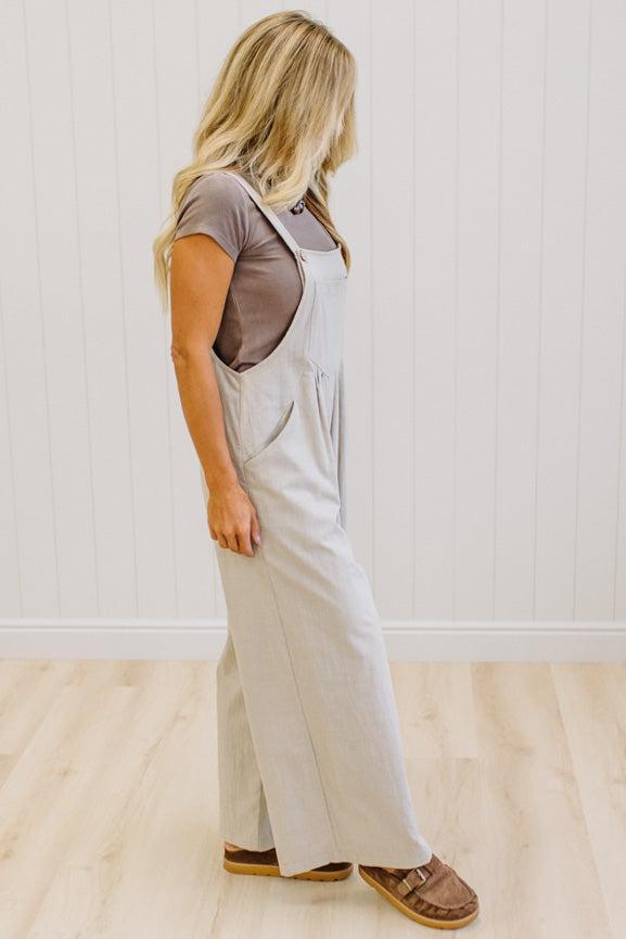 A woman modeling a pair of wide-leg stone colored utility jumpsuit over a brown t-shirt, standing in a room with light wood floors and white paneled walls.