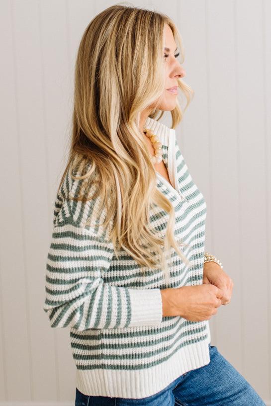 BLONDE WOMAN IN DARK JEANS STANDING IN A BRIGHTLY LIT STUDIO WITH LIGHT WOOD FLOORS AND A PANELED BACKDROP. SHE IS WEARING A CHUNKY NECKLACE AND QUARTER ZIP STRIPES SWEATER IN CREAM AND GREEN.
