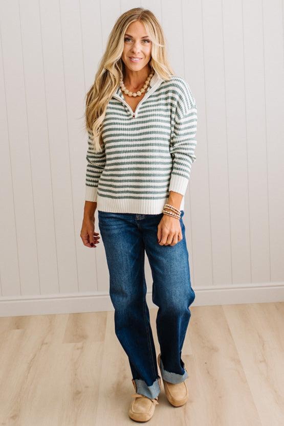 BLONDE WOMAN IN DARK JEANS STANDING IN A BRIGHTLY LIT STUDIO WITH LIGHT WOOD FLOORS AND A PANELED BACKDROP. SHE IS WEARING A CHUNKY NECKLACE AND QUARTER ZIP STRIPES SWEATER IN CREAM AND GREEN.