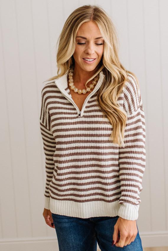 BLONDE WOMAN IN DARK JEANS STANDING IN A BRIGHTLY LIT STUDIO WITH LIGHT WOOD FLOORS AND A PANELED BACKDROP. SHE IS WEARING A CHUNKY NECKLACE AND QUARTER ZIP STRIPES SWEATER IN CREAM AND MOCHA.