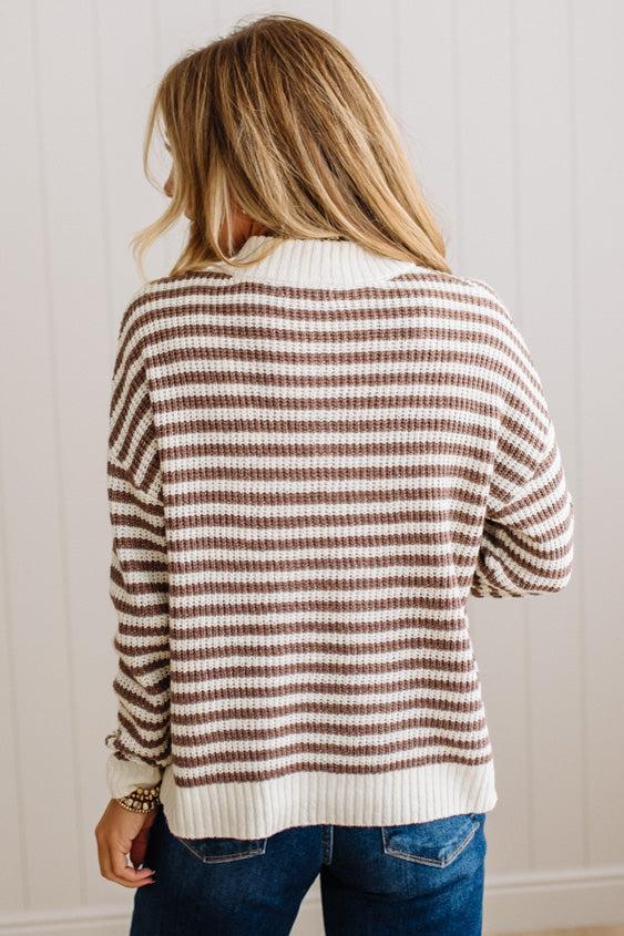 BLONDE WOMAN IN DARK JEANS STANDING IN A BRIGHTLY LIT STUDIO WITH LIGHT WOOD FLOORS AND A PANELED BACKDROP. SHE IS WEARING A CHUNKY NECKLACE AND QUARTER ZIP STRIPES SWEATER IN CREAM AND MOCHA.