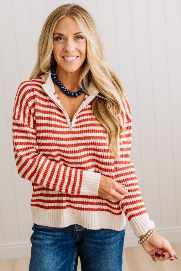 BLONDE WOMAN IN DARK JEANS STANDING IN A BRIGHTLY LIT STUDIO WITH LIGHT WOOD FLOORS AND A PANELED BACKDROP. SHE IS WEARING A CHUNKY NECKLACE AND QUARTER ZIP STRIPES SWEATER IN CREAM AND RUST.
