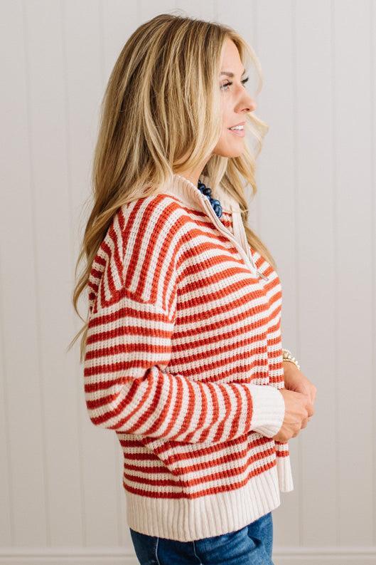 BLONDE WOMAN IN DARK JEANS STANDING IN A BRIGHTLY LIT STUDIO WITH LIGHT WOOD FLOORS AND A PANELED BACKDROP. SHE IS WEARING A CHUNKY NECKLACE AND QUARTER ZIP STRIPES SWEATER IN CREAM AND RUST.