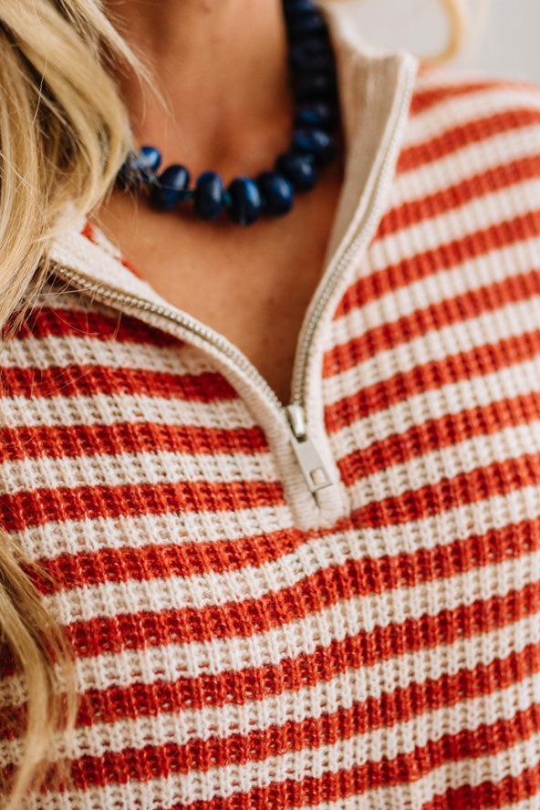 BLONDE WOMAN IN DARK JEANS STANDING IN A BRIGHTLY LIT STUDIO WITH LIGHT WOOD FLOORS AND A PANELED BACKDROP. SHE IS WEARING A CHUNKY NECKLACE AND QUARTER ZIP STRIPES SWEATER IN CREAM AND RUST.