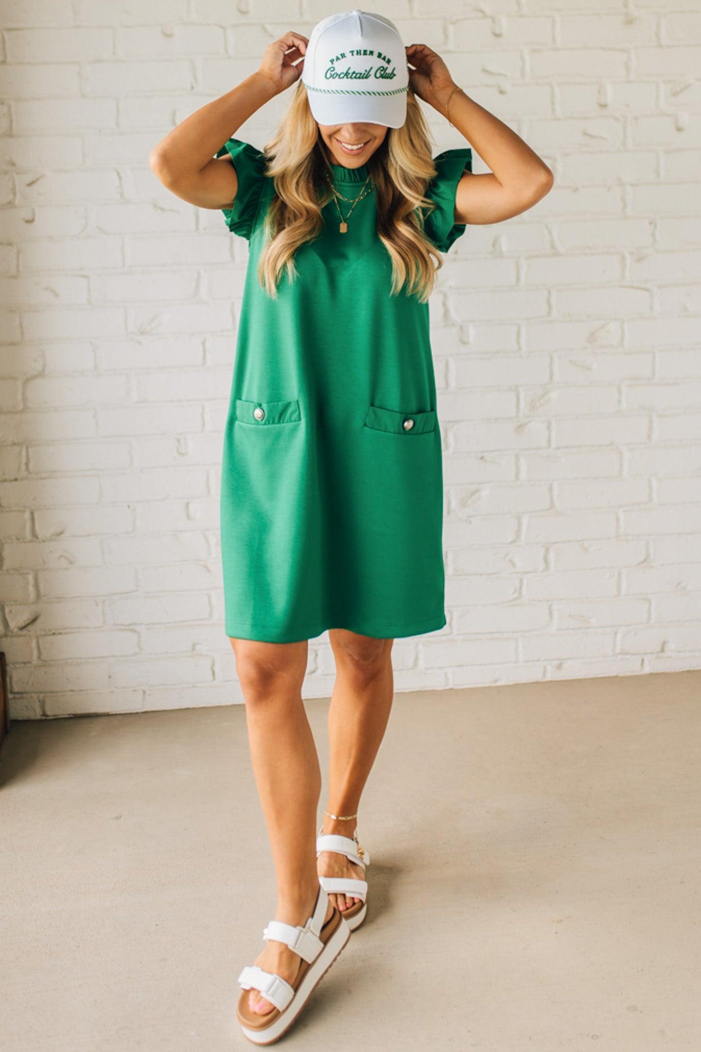 Blonde woman wearing a ruffled cap sleeves shift dress with pockets, ruffled neckline, button detail front pockets, button closure back, in sleek soft fabric.