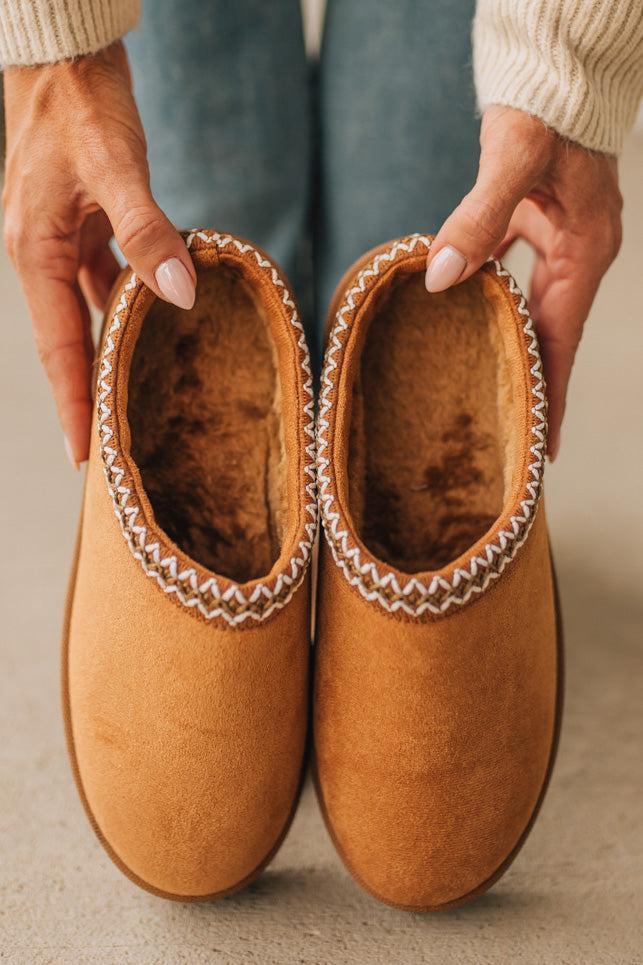 Brown slippers with decorative trim held by a person against a neutral background