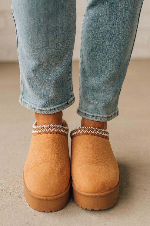 Tan slip-on shoes with patterned straps worn with blue jeans on a neutral background