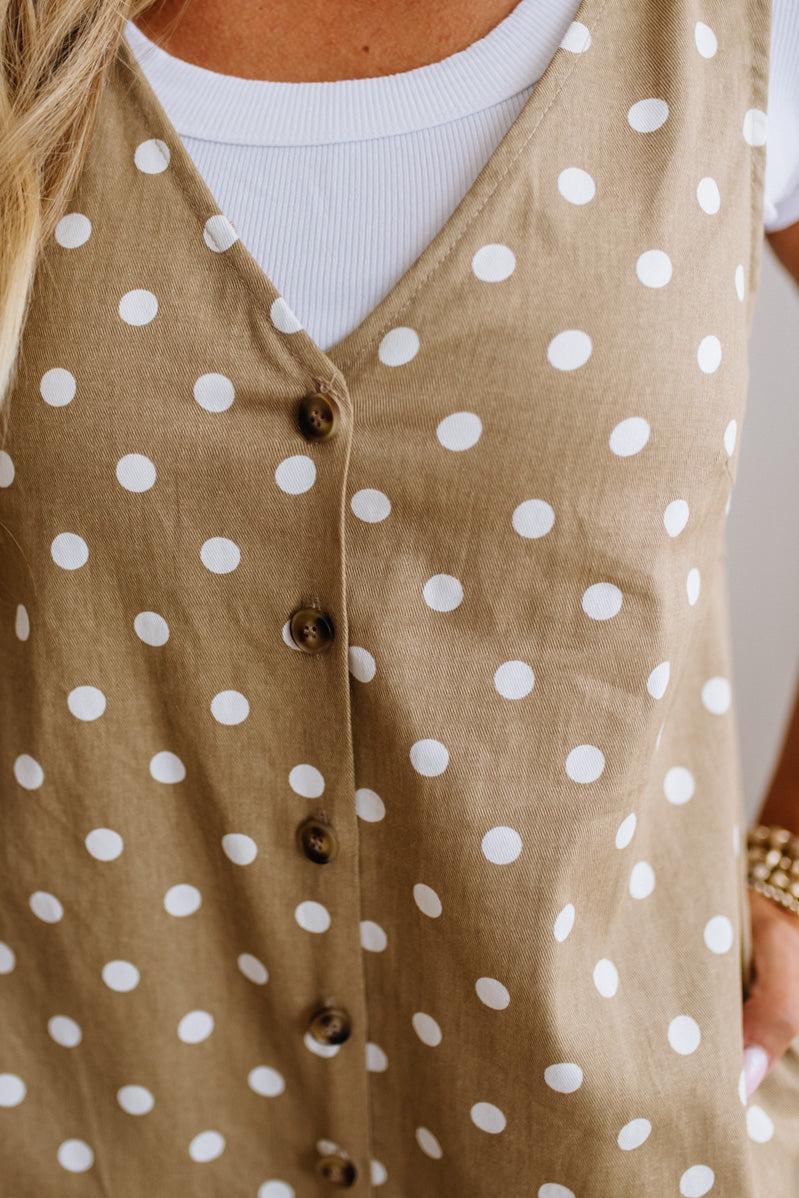 A close-up of a woman wearing a sleeveless tan vest with white polka dots and dark brown buttons over a white ribbed top.