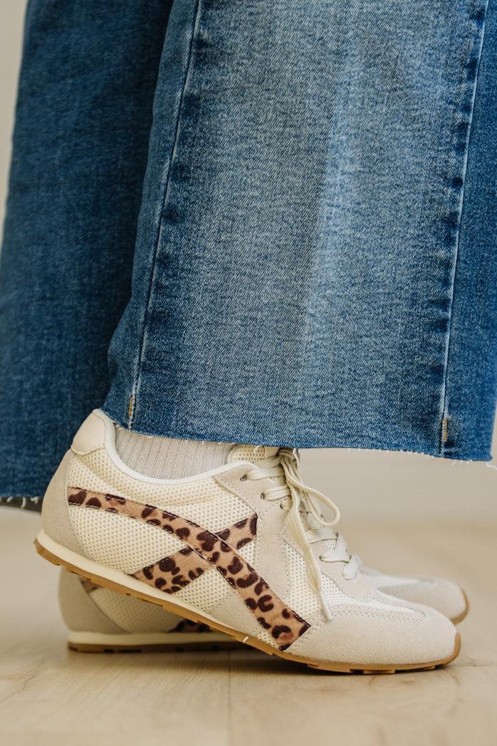 White sneakers with leopard print accents worn with blue jeans on a light wooden floor.
