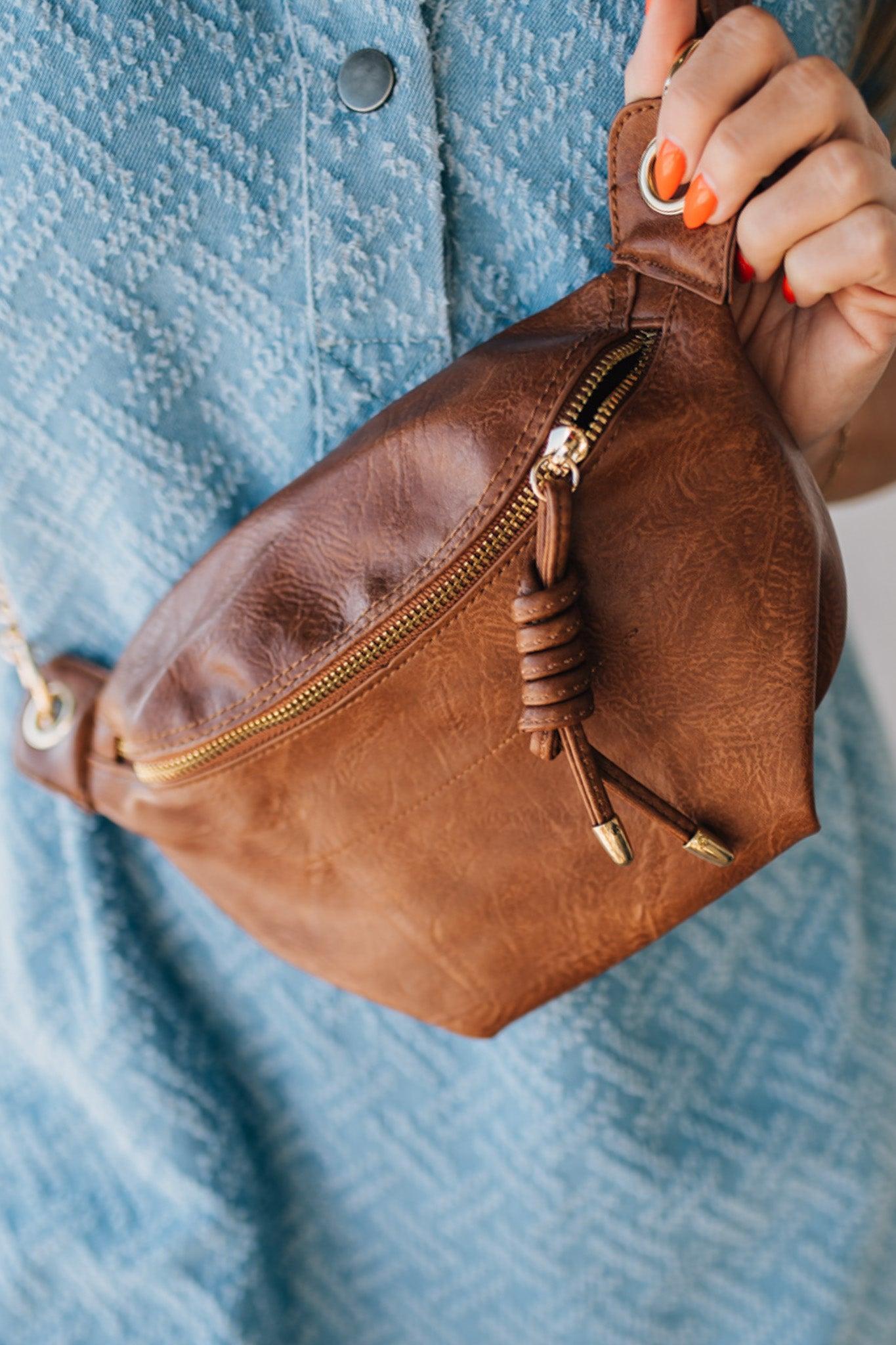 Blonde woman wearing a blue denim dress and cognac colored vegan leather crossbody back with gold hardware.
