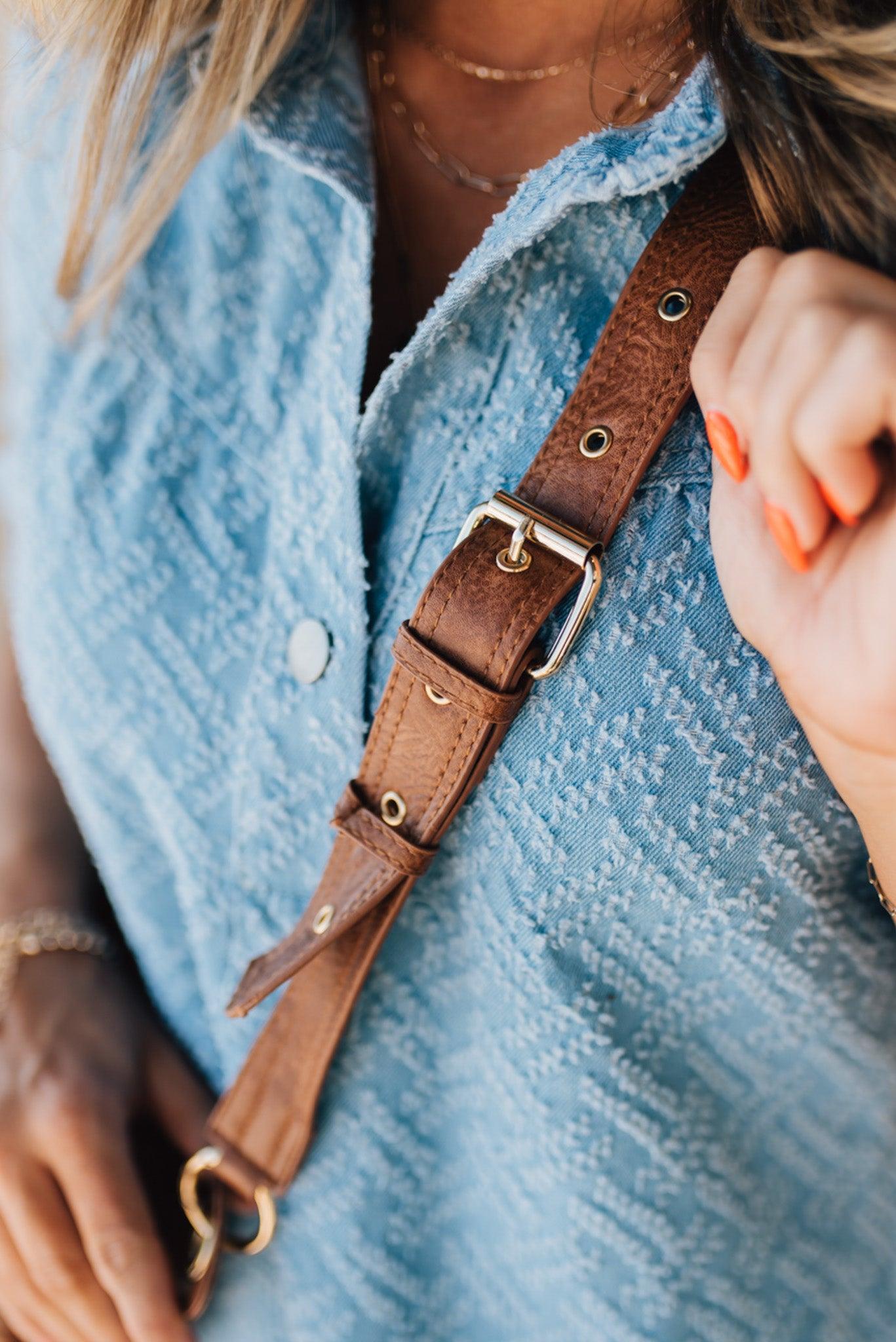 Blonde woman wearing a blue denim dress and cognac colored vegan leather crossbody back with gold hardware.