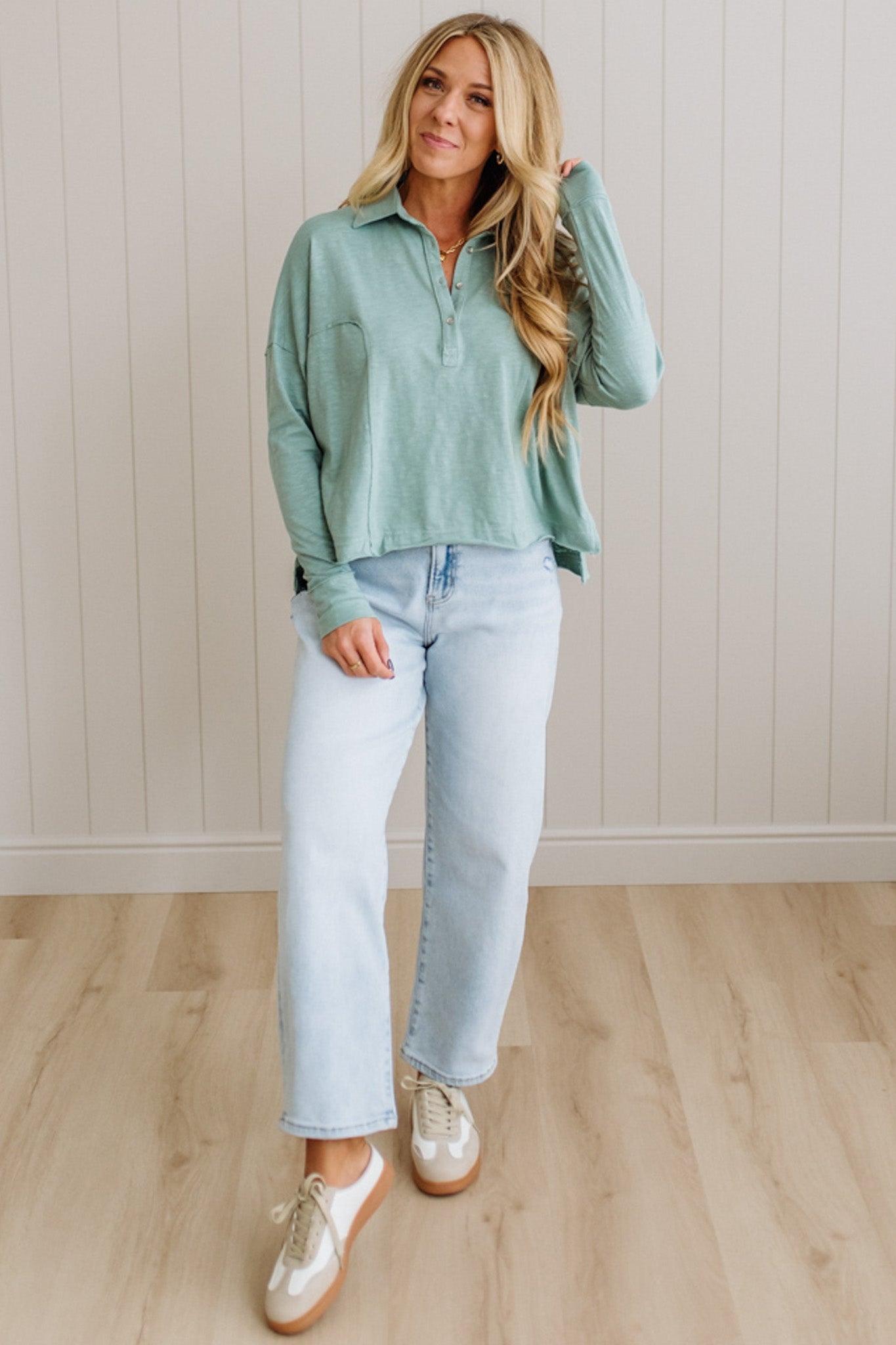 Woman wearing a light green shirt and light blue jeans standing against a white paneled wall.