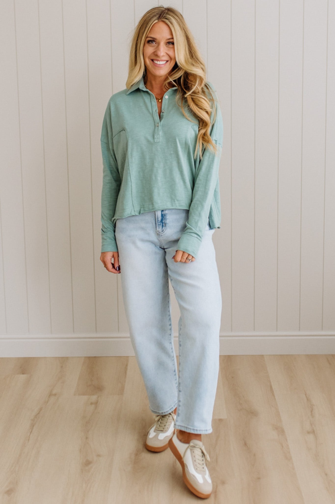 Woman wearing a green long-sleeve shirt and light blue jeans standing against a white paneled wall.