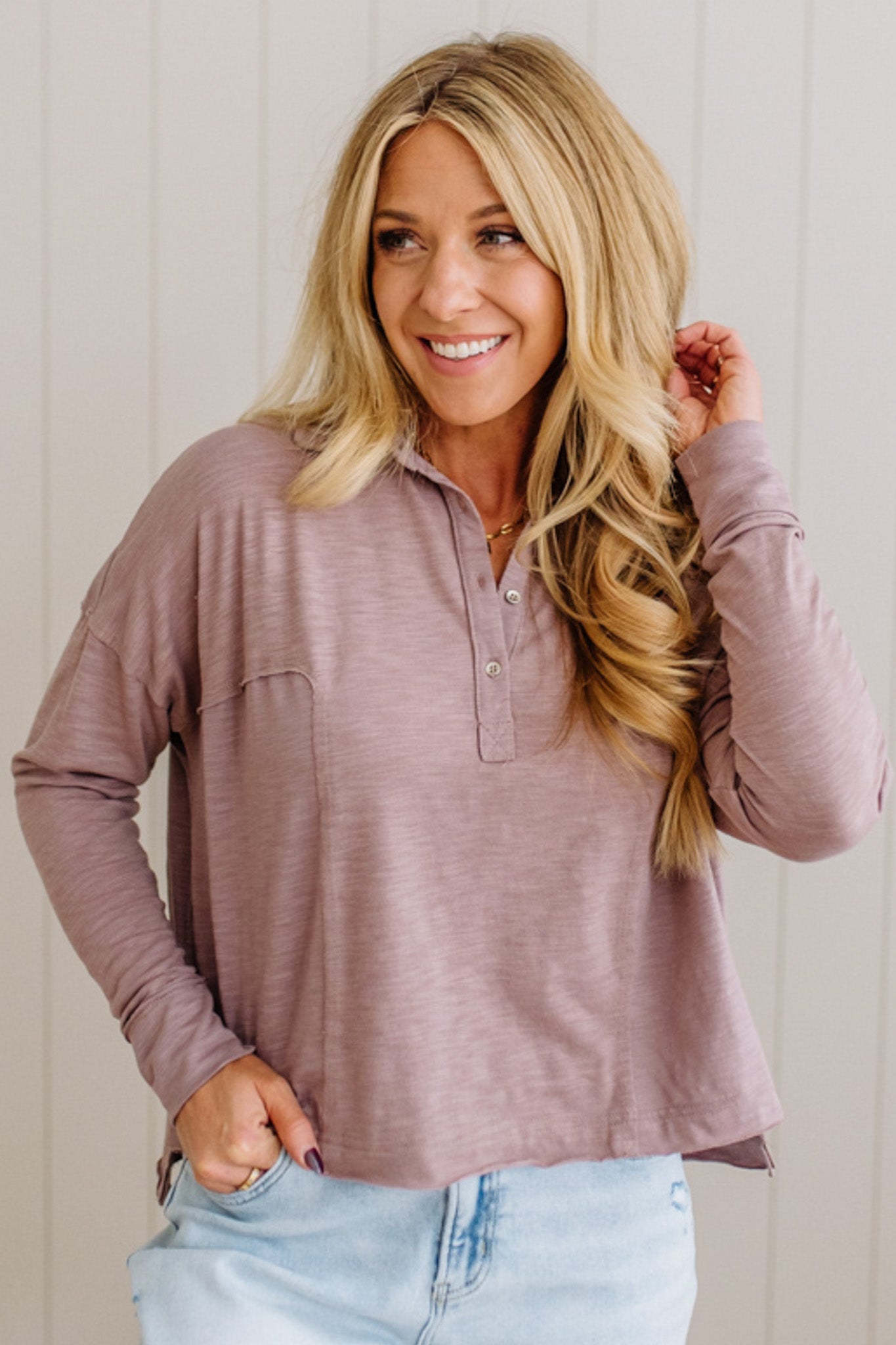 Woman wearing a mauve long-sleeve shirt and light blue jeans against a white wall.