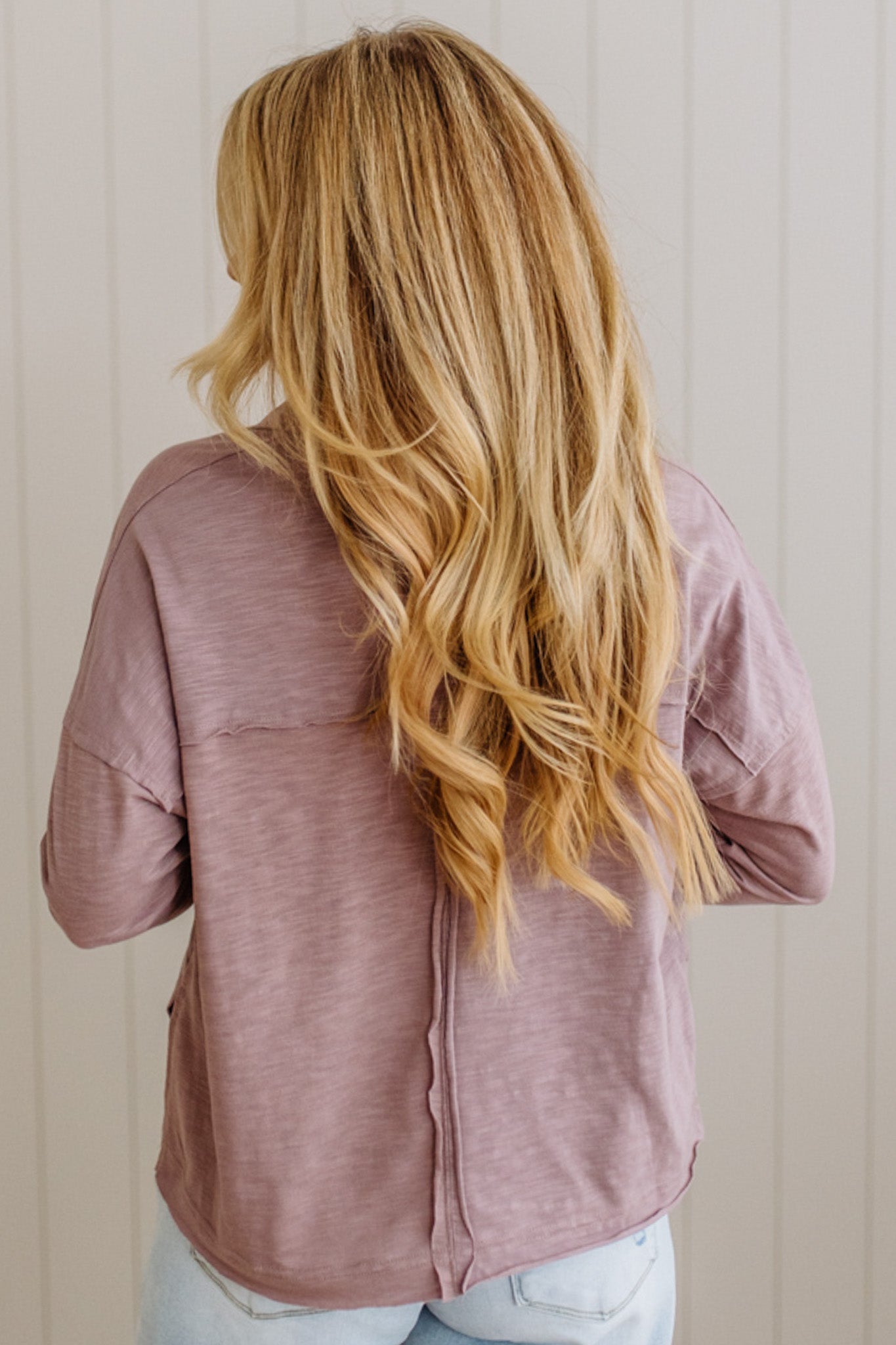 Person wearing a mauve shirt with long blonde hair, standing against a neutral background